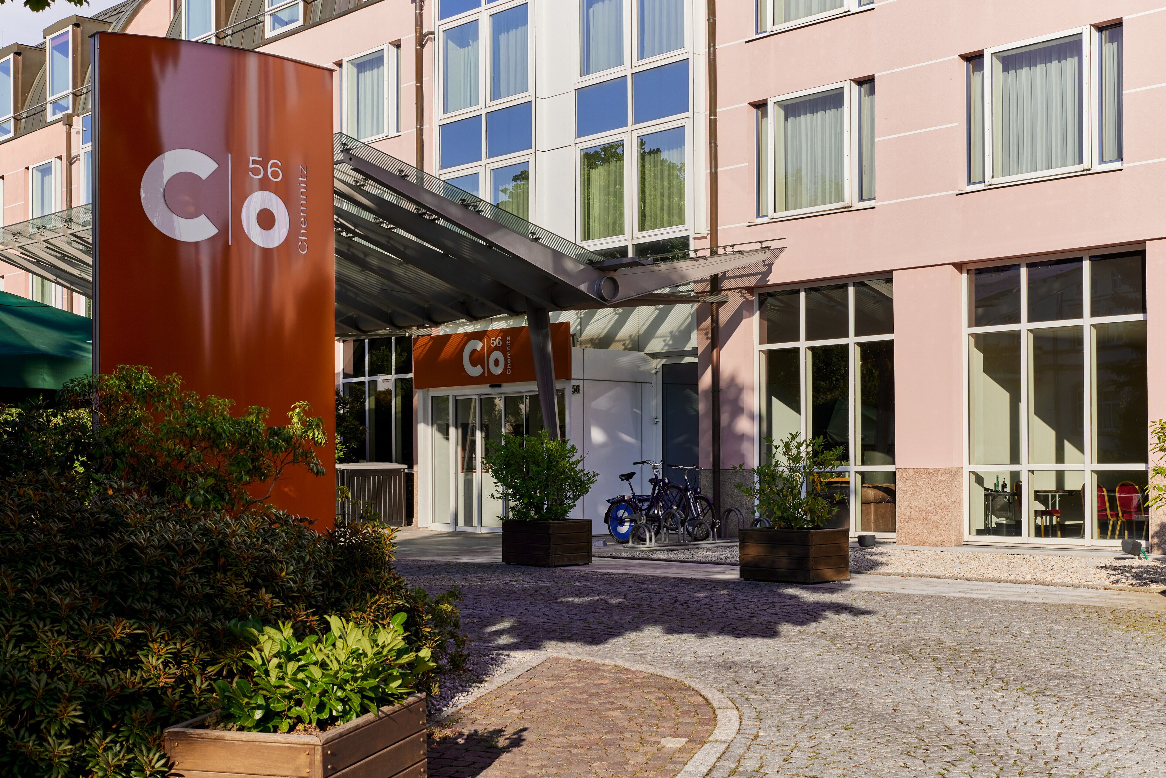 A hotel entrance with a large sign, potted plants, and bicycles parked outside.