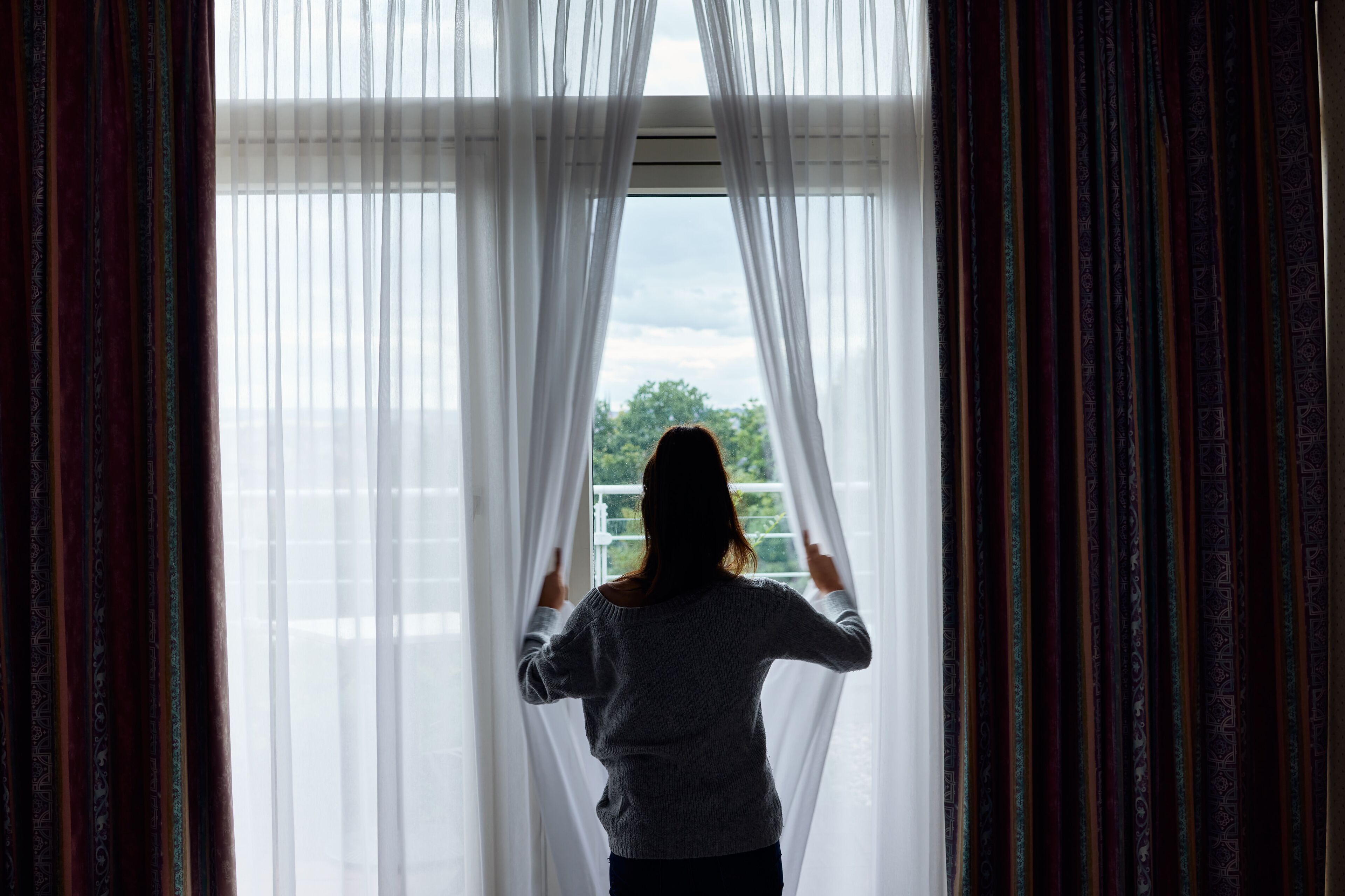 Eine Person steht vor einem Fenster mit transparenten Vorhängen und blickt hinaus.