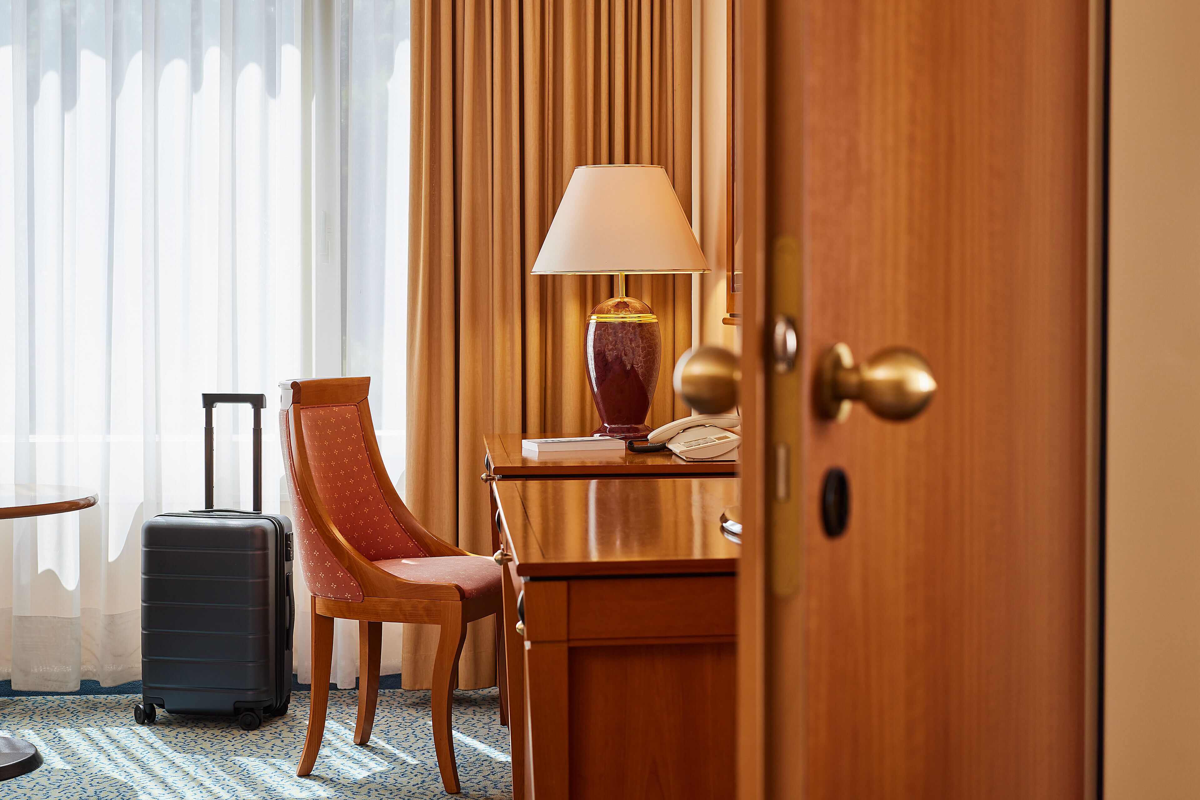 Ein Hotelzimmer mit einem Holztisch, einer Lampe, einem Stuhl, einem Koffer und einer Tür.