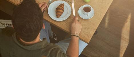 Una persona sentada en una mesa de madera con un croissant, una taza de café y un vaso de jugo.