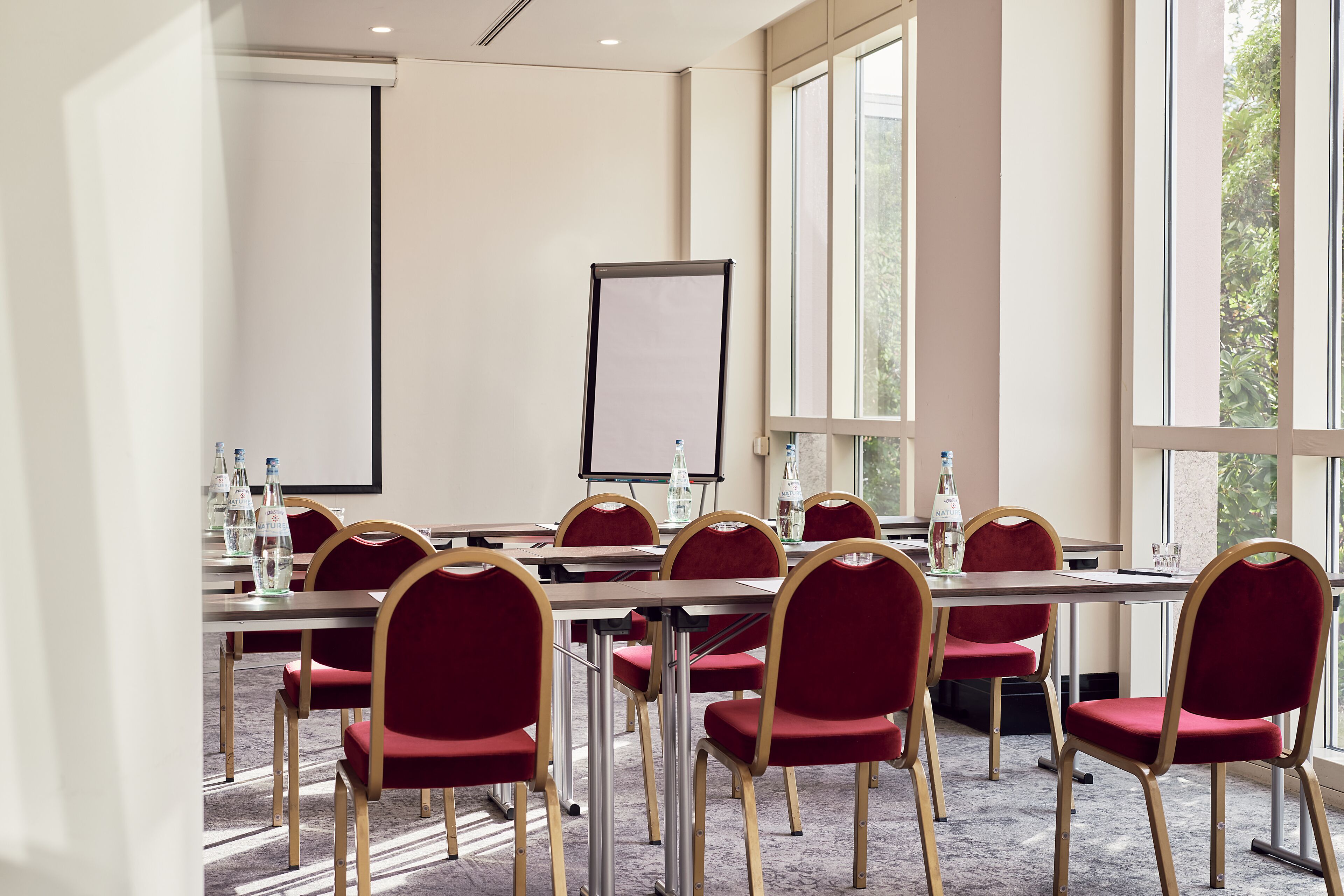 Ein Konferenzraum mit langem Tisch, roten Stühlen, einer Projektionsleinwand und großen Fenstern.