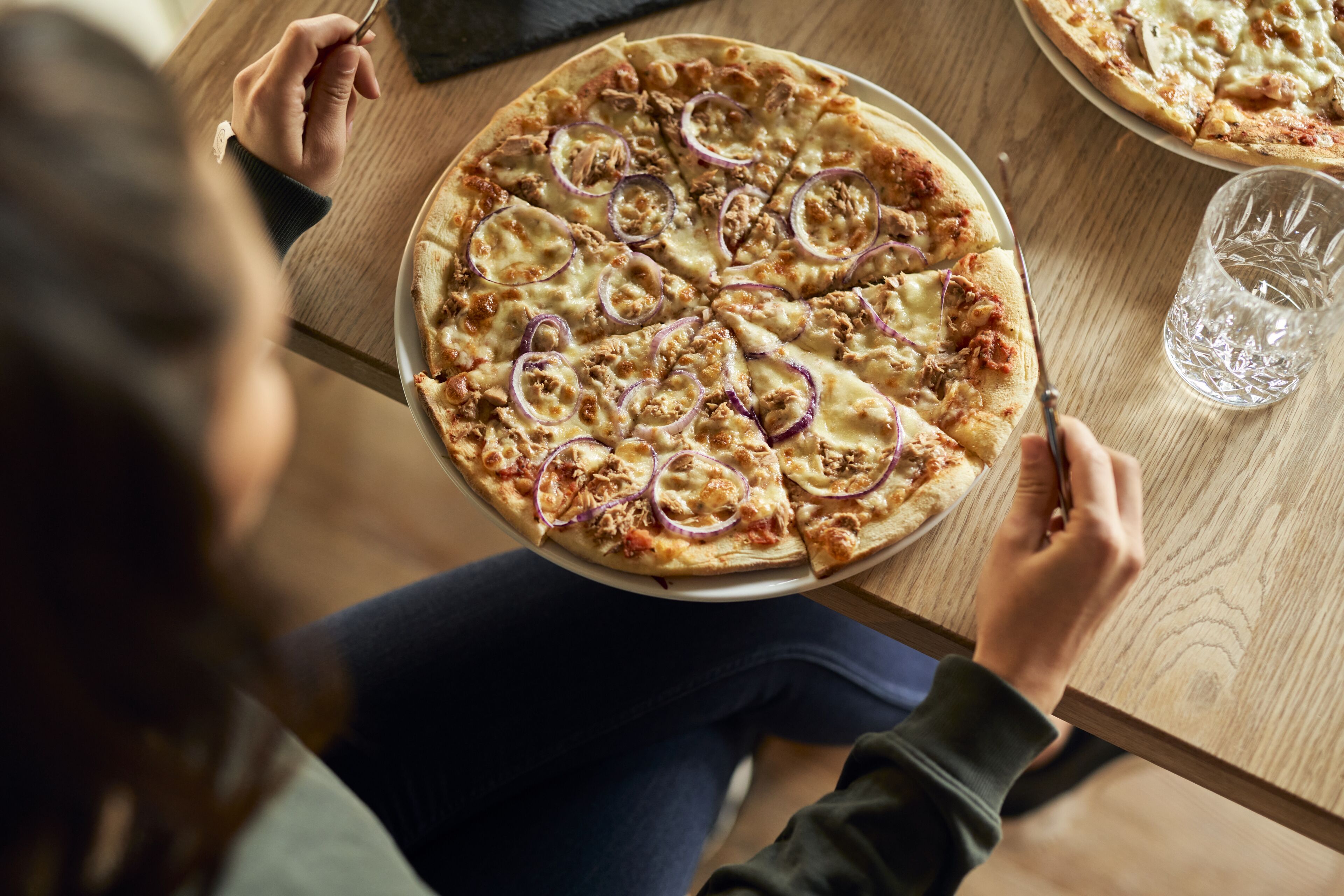 Eine Person sitzt an einem Holztisch mit einer Pizza und einem Glas Wasser.