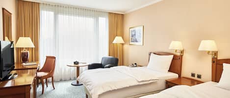 A hotel room with two beds, a desk with a TV, a chair, a small table, and a window with curtains.