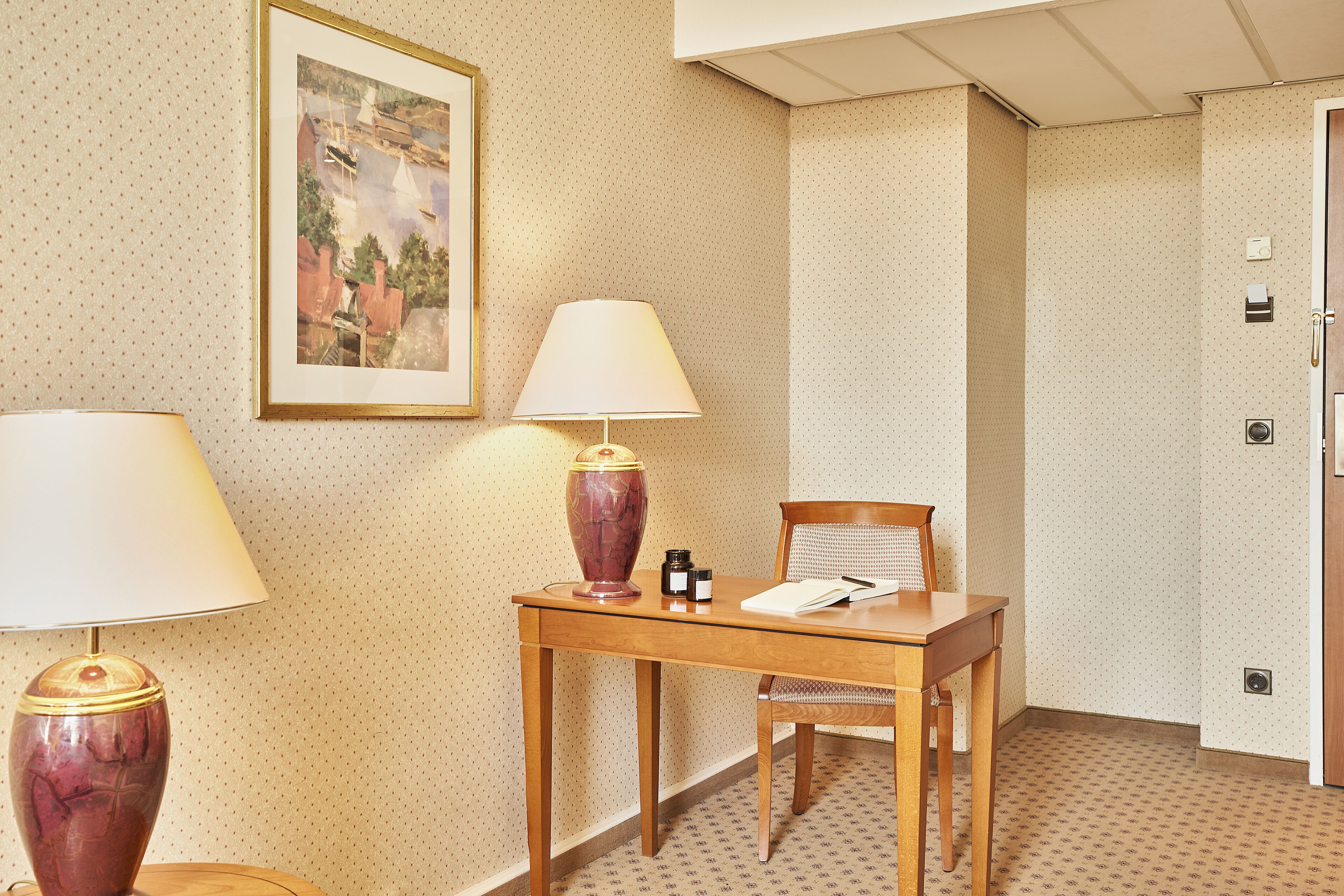 Habitación de hotel con un escritorio de madera, dos lámparas, una silla, un cuadro en la pared y una puerta con manija.