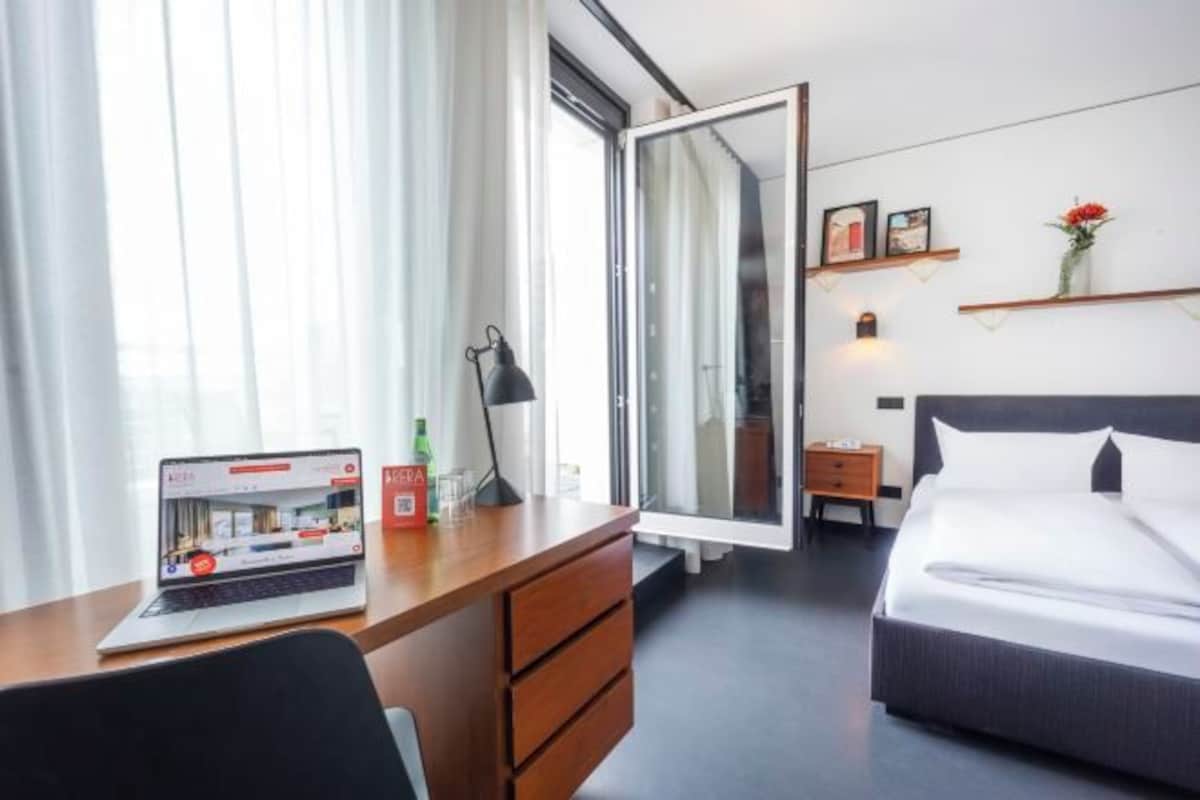 A hotel room with a desk, laptop, bed, bedside table, lamp, and a window with curtains.
