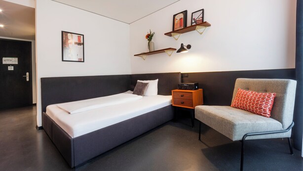 Una habitación de hotel moderna con cama, una silla, una mesita de noche y estantes en la pared.