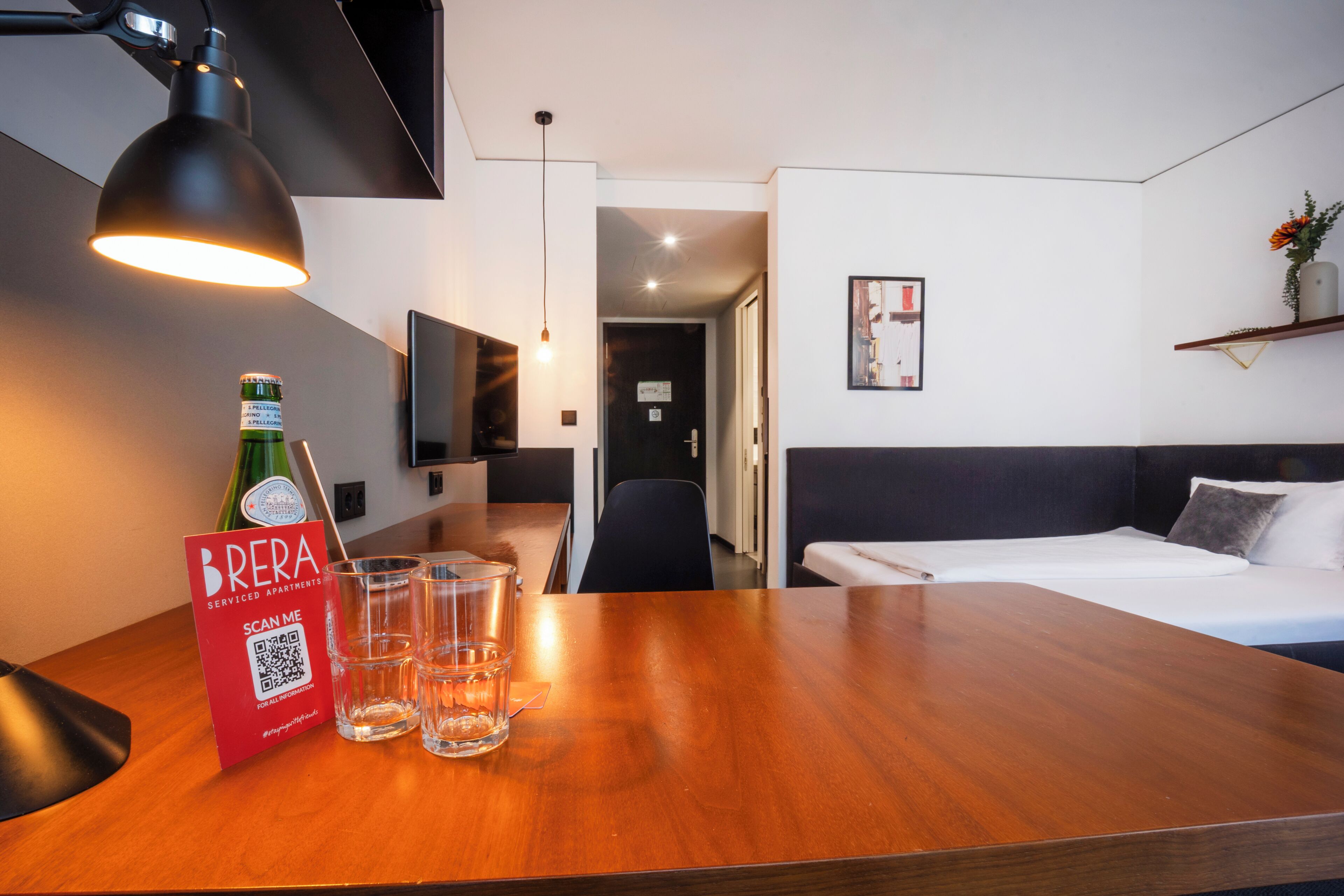 A hotel room with a wooden table, a bed, a TV, and a QR code sign.