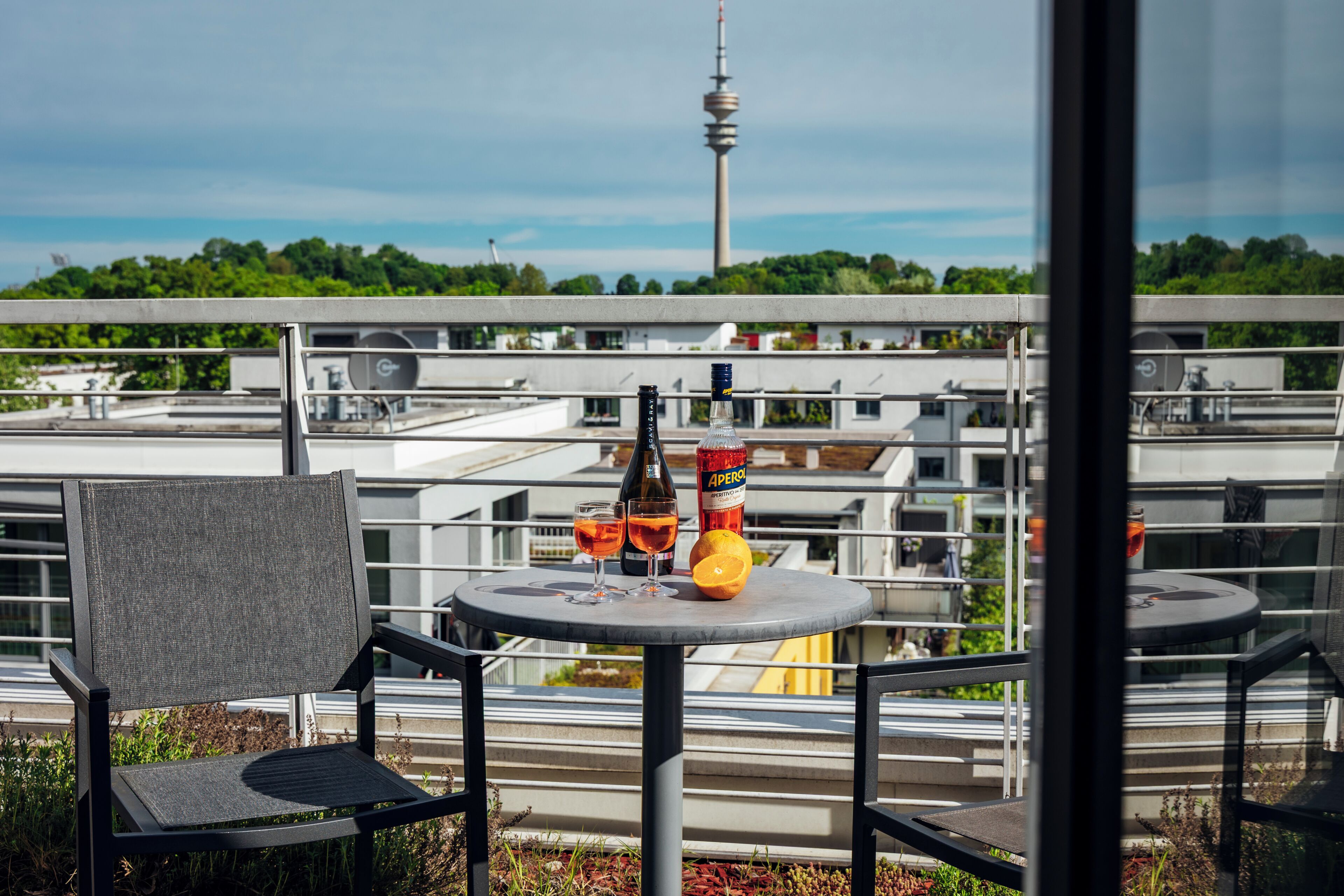 Un balcon avec une table, des chaises, une bouteille et des verres, donnant sur un paysage urbain avec des immeubles et une tour.