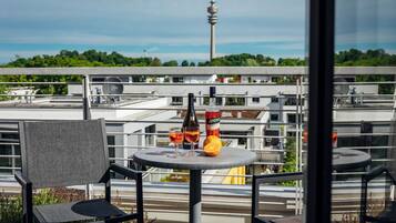 Un balcon avec une table, des chaises, une bouteille et des verres, donnant sur un paysage urbain avec des immeubles et une tour.