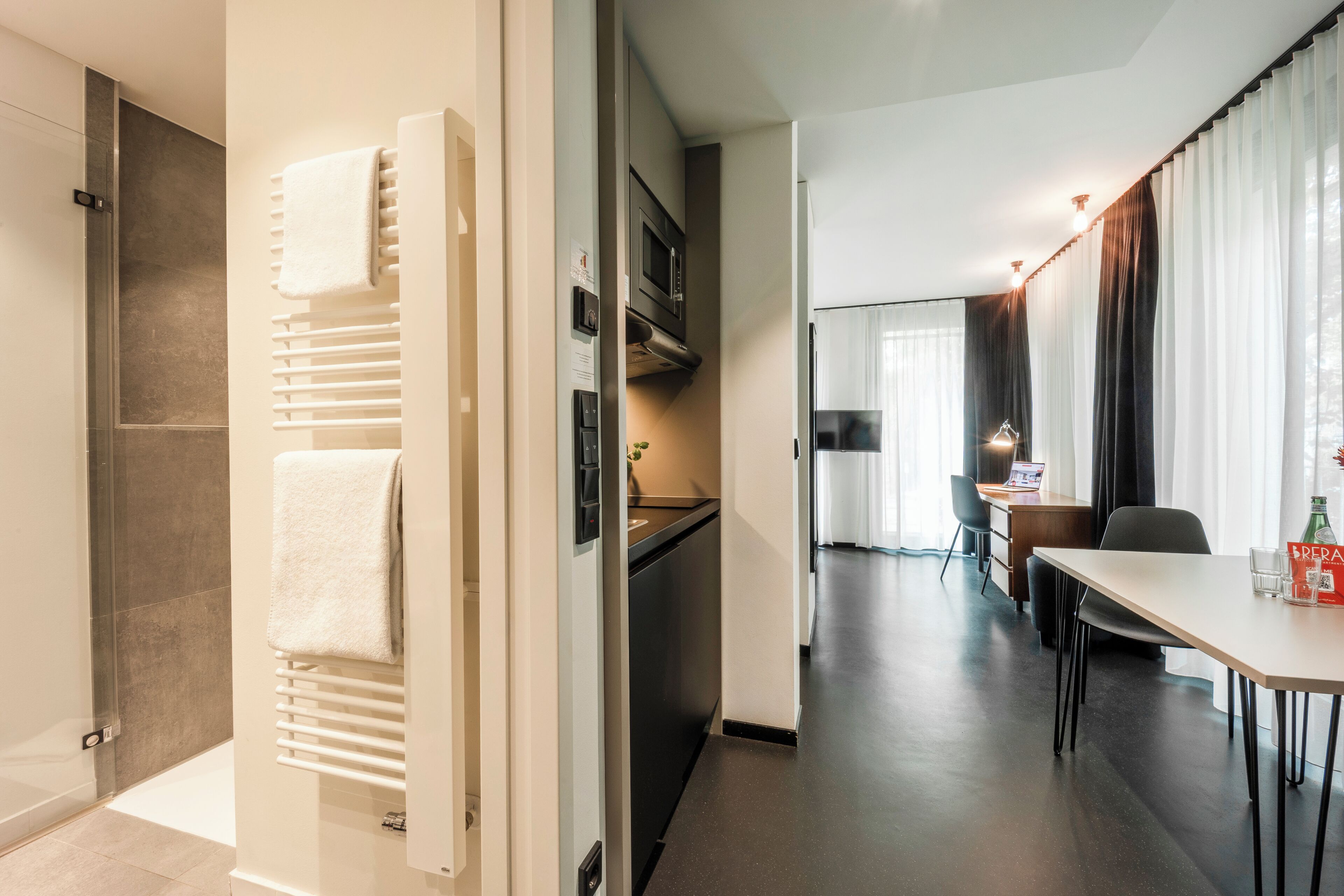 Habitación de hotel moderna con ducha, toallero, zona de comedor con mesa y sillas, y cocina con refrigerador y microondas.