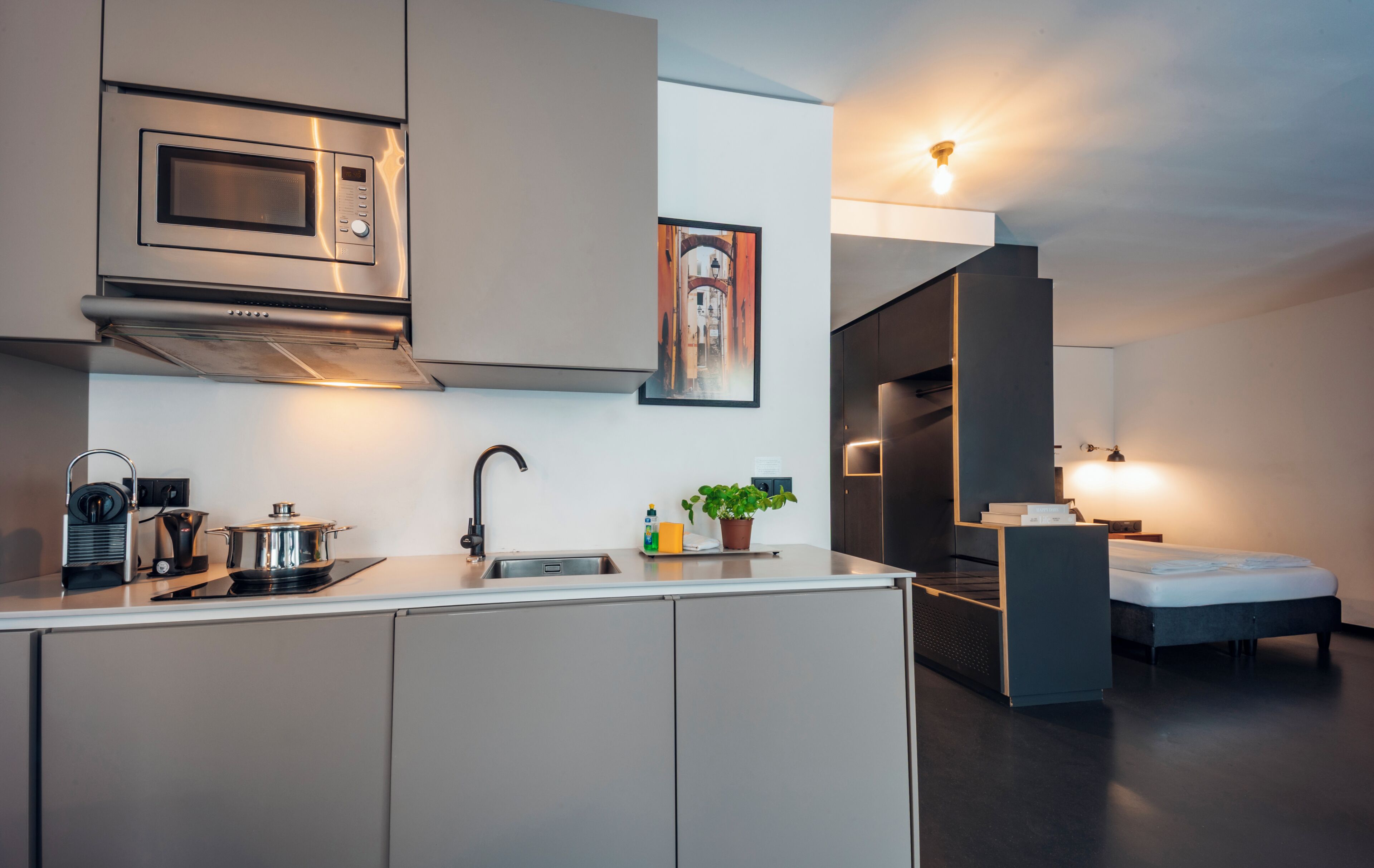 A modern kitchen with a built-in microwave, a kettle, and a sink. There is a small dining area with a bed and a nightstand.