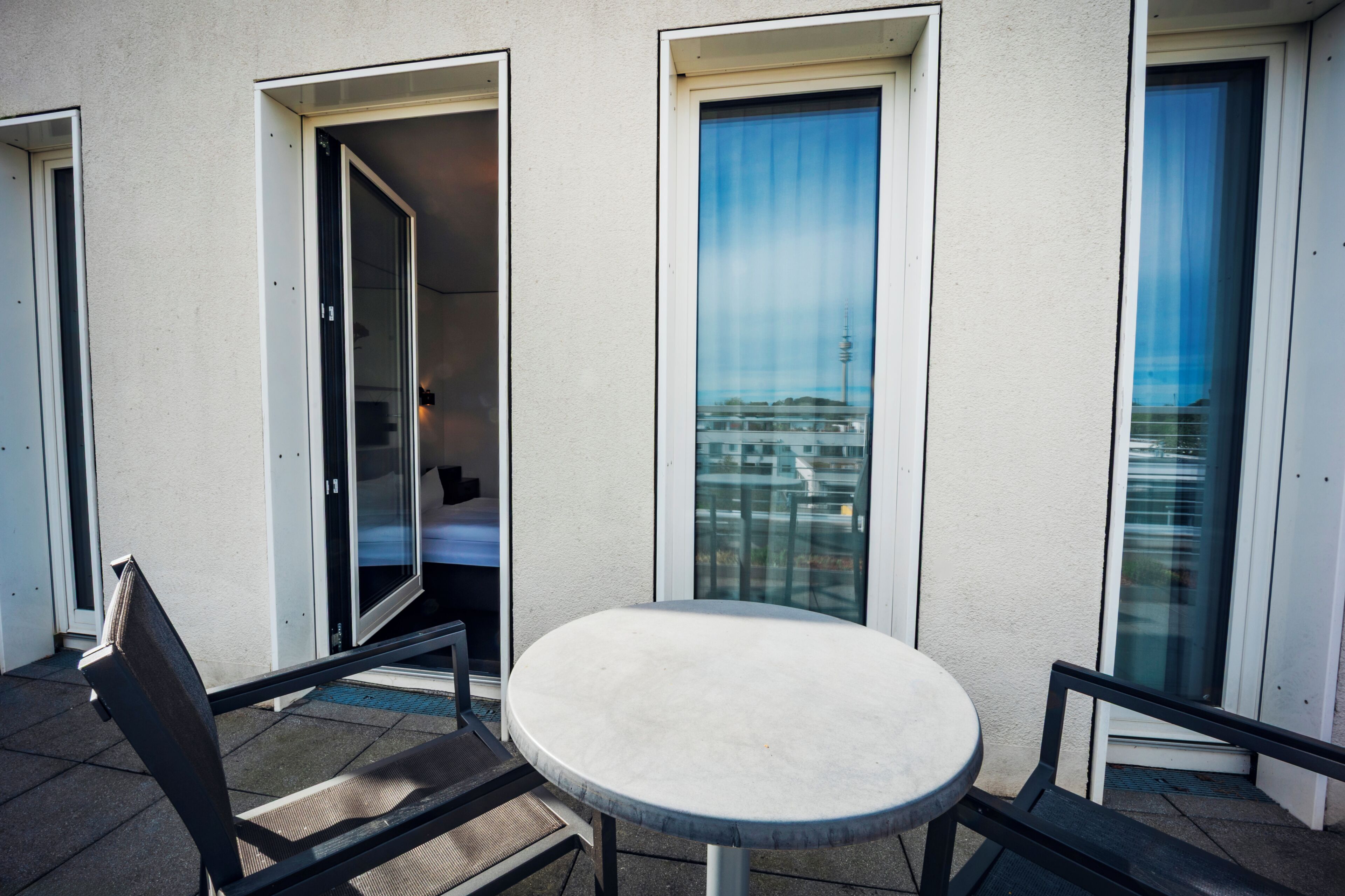 Ein Balkon mit rundem Tisch und Stühlen, eine Tür zu einem Innenraum sowie der Blick auf ein Gebäude mit einem Turm.