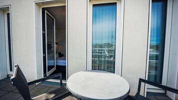 Ein Balkon mit rundem Tisch und Stühlen, eine Tür zu einem Innenraum sowie der Blick auf ein Gebäude mit einem Turm.