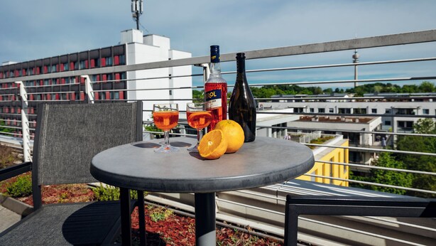 A table with a bottle, two glasses, and an orange on a balcony overlooking a cityscape.