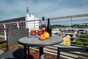 A table with a bottle, two glasses, and an orange on a balcony overlooking a cityscape. - Brera Serviced Apartments Schwabing (Munich)