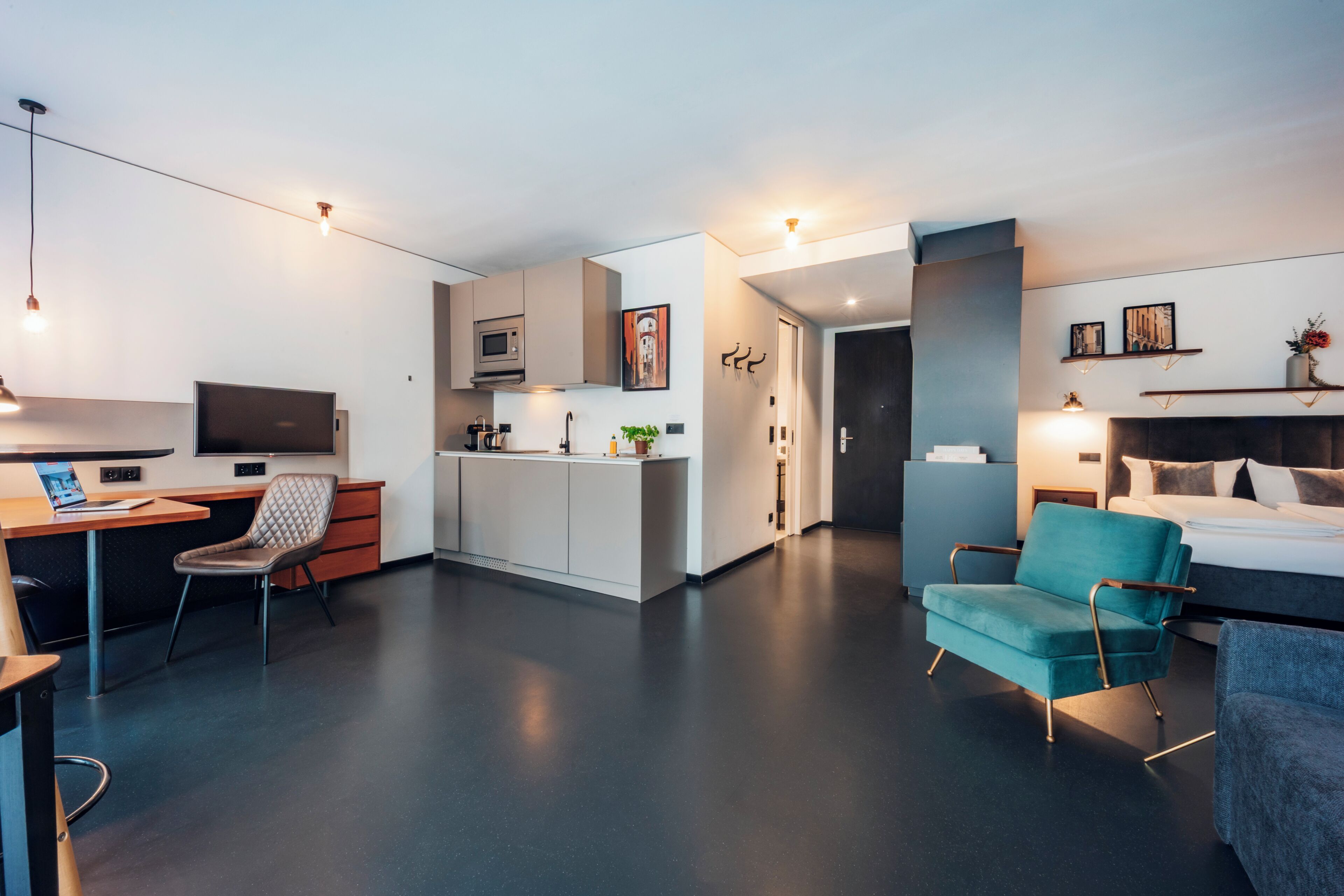 A modern hotel room with a kitchenette, a desk with a laptop, a comfortable seating area, and a bed.