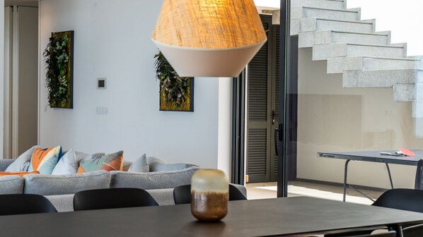 A modern living room with a grey sofa, colorful cushions, a dining table with chairs, a pendant lamp, and a staircase.