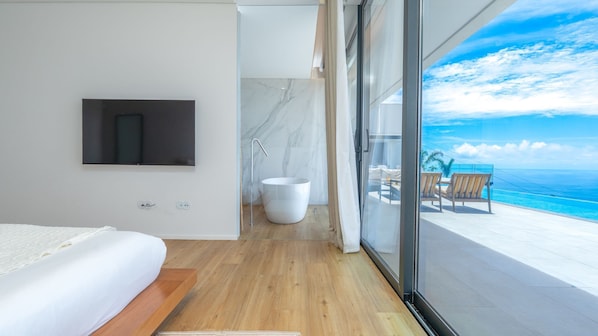 A modern bedroom with a large bed, a mounted TV, a bathtub, and a balcony with a view of the ocean.