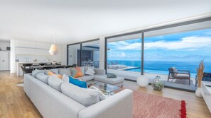 Un salon moderne avec un grand canapé gris, une table basse en verre et une vue sur l’océan.