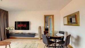 Un salon moderne avec un téléviseur à écran plat affichant le logo « NETFLIX », un coin repas avec une table ronde et des chaises, et un grand miroir sur le mur.