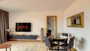 Una sala de estar moderna con un televisor de pantalla plana mostrando 'NETFLIX', un comedor con una mesa redonda y sillas, y un espejo grande en la pared.