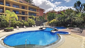 A swimming pool with a wooden deck and lounge chairs, surrounded by tropical plants and a multi-story building in the background.