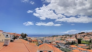 Un paysage urbain côtier avec des bâtiments aux toits de tuiles rouges, un ciel bleu dégagé et la mer à l’horizon.