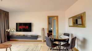 A modern living room with a flat-screen TV displaying 'NETFLIX', a dining area with a round table and chairs, and a large mirror on the wall.