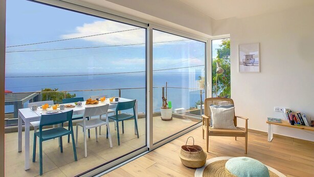 Une chambre moderne dotée d’une grande porte-fenêtre coulissante donnant sur un balcon avec vue sur la mer. On y trouve une table à manger avec des chaises, un fauteuil en osier et une petite bibliothèque.