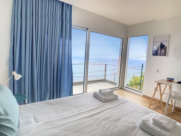 A hotel room with a large bed, a desk, and a view of the sea through large windows. - Villa Omar by LovelyStay (São Tiago)
