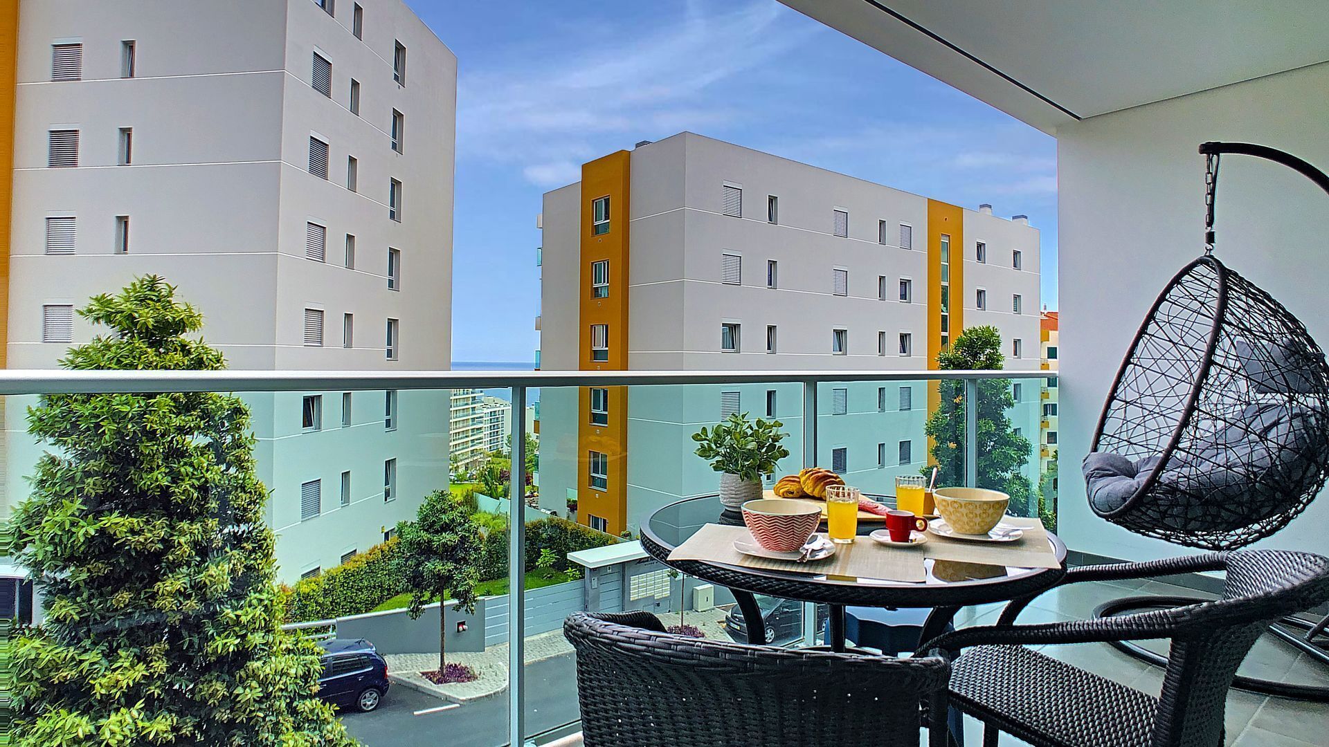 A balcony with a glass railing, wicker furniture, a hanging chair, and a table with breakfast items.