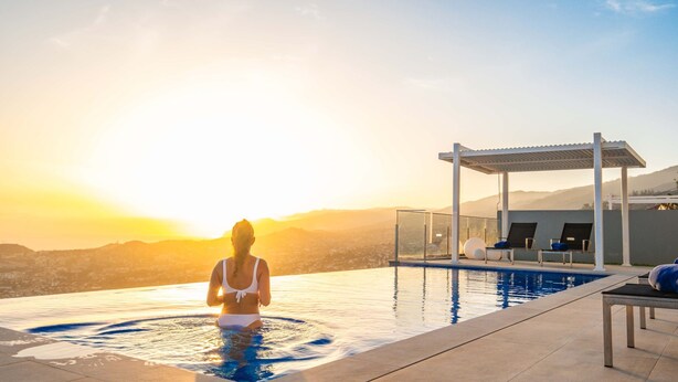 A person in a white bikini in a pool with a sunset view, lounge chairs, and a pergola.