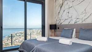 A hotel room with a large window offering a city view, a bed with two pillows, and a nightstand with a lamp.