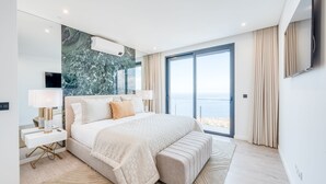 Un dormitori modern amb un llit gran, una tauleta de nit, una televisió i vista al mar a través d'una porta corredissa de vidre.