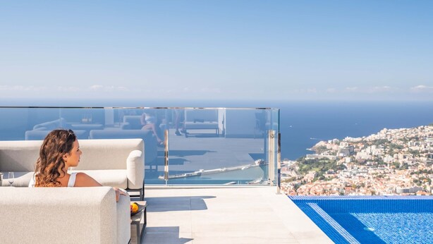Una piscina d'hidromassatge sense arestes envuelta en vidre amb vista a la ciutat, una zona de relaxació amb seients i un cel serè.
