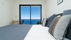 A bedroom with a large bed, a view of the ocean through a sliding door, and a small chair by the door. - Evoque Villa by LovelyStay (Arco Da Calheta)