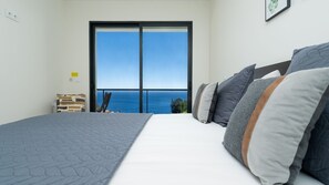 A bedroom with a large bed, a view of the ocean through a sliding door, and a small chair by the door. - Evoque Villa by LovelyStay (Arco Da Calheta)