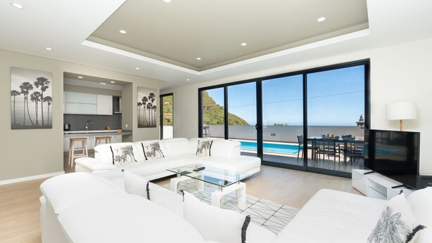 A modern living room with a sofa, a coffee table, and a TV. There is a kitchen in the background and a large window with a view of the sea. - Evoque Villa by LovelyStay (Arco Da Calheta)