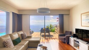 Un salon moderne avec un canapé, un fauteuil et un meuble pour la télévision. On aperçoit un balcon avec une table et des chaises, d’où l’on peut admirer un paysage magnifique.