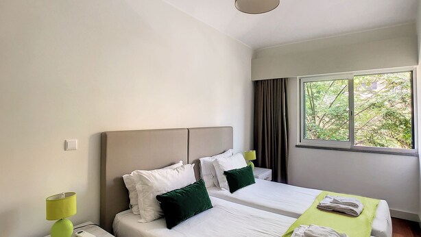 Une chambre d’hôtel avec deux lits, une fenêtre donnant sur un espace verdoyant et une table de chevet avec une lampe.
