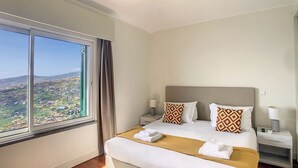 Una habitación de hotel con un ventanal grande, una cama con sábanas blancas, dos almohadas estampadas, un cabecero de madera y una mesita de noche con una lámpara.