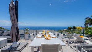 Une table à manger avec vue sur l’océan, des sièges d’extérieur et un espace abrité avec un parasol.