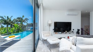 A modern living room with a large TV, white furniture, and a view of a swimming pool.