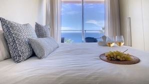 Habitación de hotel con una cama grande, dos almohadas, una bandeja con uvas y copas de vino, y vista al océano a través de una puerta corredera.