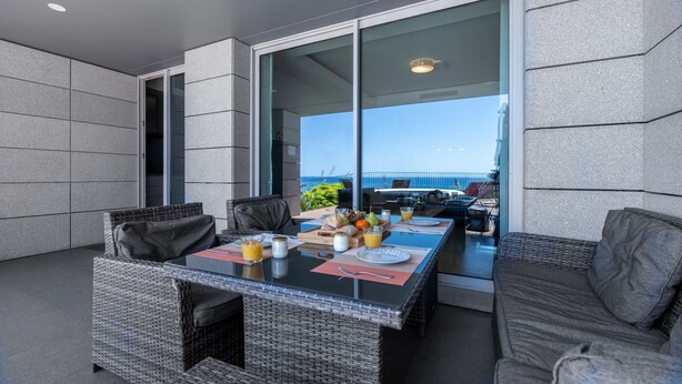 A dining area with wicker furniture, a table set with plates, glasses, and a fruit tray, and a view of the sea through large glass doors. - Over the Cliff Apartment by LovelyStay (Funchal)