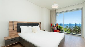 A bedroom with a large bed, a wooden headboard, a nightstand, a bedside table, and a view of the ocean through a balcony.