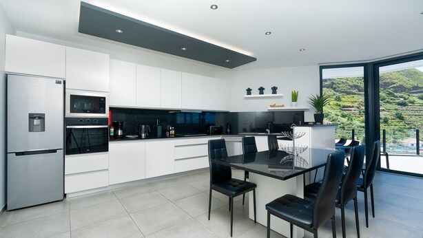 A modern kitchen with white cabinetry, a built-in oven, a refrigerator, and a dining area with a glass table and chairs. - Villa Stella by LovelyStay (Arco Da Calheta)