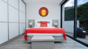A modern bedroom with a large bed, a bench, and a view of greenery through a large window. - Villa Stella by LovelyStay (Arco Da Calheta)