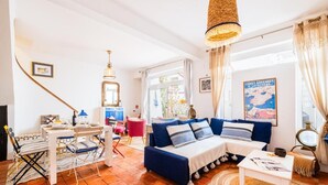 A cozy living room with a sofa, dining table, and chairs. There is a staircase leading to another room, and a large window with curtains. - Fisherman's cottage a few steps from the beach (Olhos de Água)