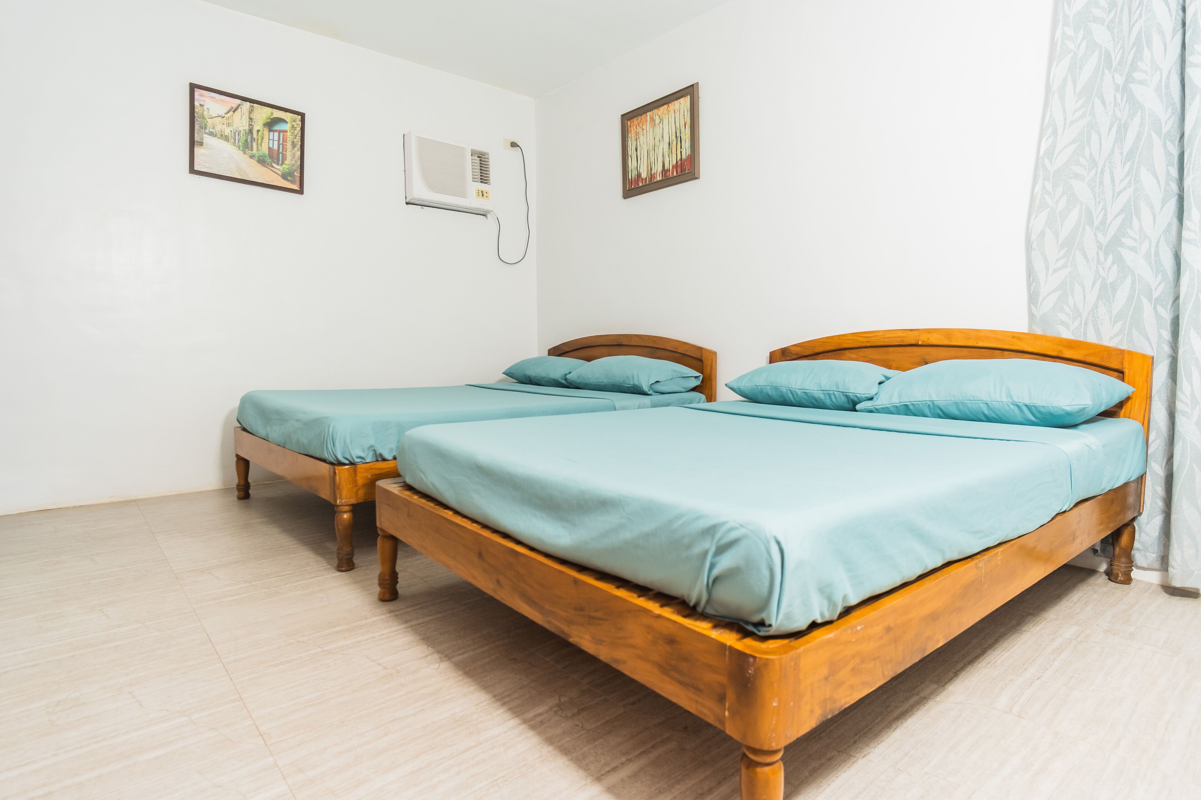 A room with two wooden beds, each with a light blue mattress and matching pillows. There are two framed pictures on the walls, a white air conditioning unit, and a window with a white curtain.